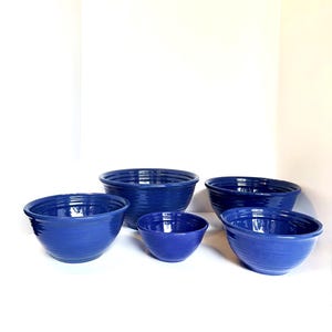 May include: A collection of five cobalt blue ceramic bowls of varying sizes. Each bowl has a ribbed texture and a rounded shape, suitable for serving or food preparation. The bowls are arranged on a white surface.