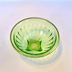 May include: A light green glass bowl with a square base and a round rim. The bowl has vertical ridges that radiate from the base to the rim. The bowl is made of a translucent glass material.