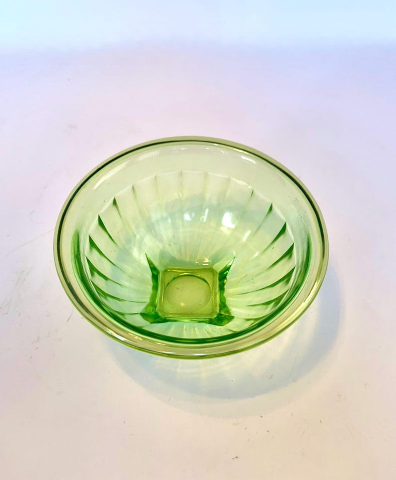 May include: A light green glass bowl with a square base and a round rim. The bowl has vertical ridges that radiate from the base to the rim. The bowl is made of a translucent glass material.