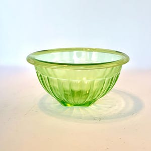 May include: A small, round, light green glass bowl. The bowl has a ribbed exterior design and a smooth, rounded rim. The glass is translucent, allowing light to pass through. The bowl is likely used for serving or mixing.