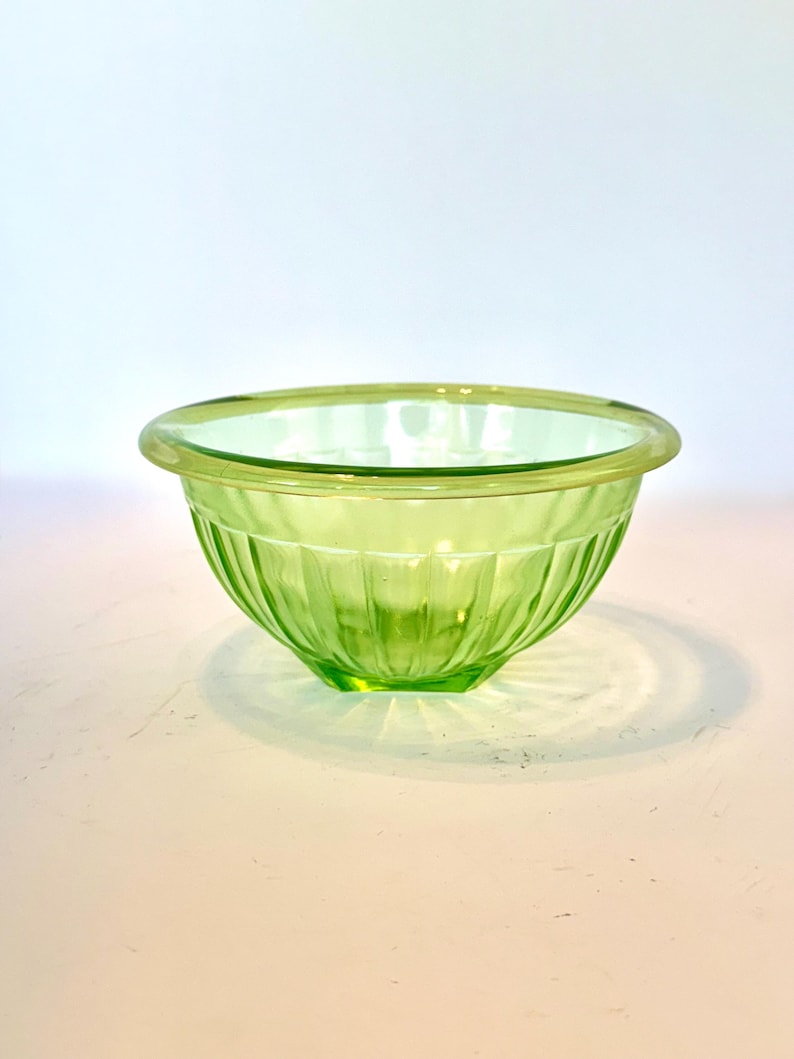May include: A small, round, light green glass bowl. The bowl has a ribbed exterior design and a smooth, rounded rim. The glass is translucent, allowing light to pass through. The bowl is likely used for serving or mixing.