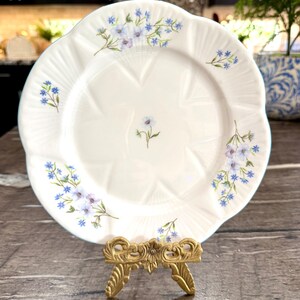 May include: White porcelain plate with a scalloped edge, adorned with delicate blue and purple floral sprigs. The plate is displayed on a decorative gold-colored stand, showcasing its elegant design. A charming piece for serving or display.