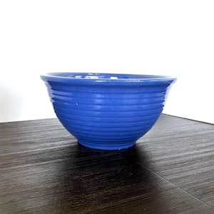 May include: A vibrant blue ceramic bowl with a ribbed exterior. The bowl has a rounded shape with a slightly flared rim. The bowl is sitting on a dark wooden surface. This bowl is suitable for serving food.