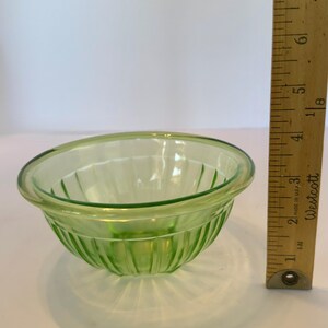May include: A small, light green glass bowl with vertical ribbed detailing. The bowl has a rounded shape with a slightly flared rim. A wooden ruler is positioned next to the bowl, showing measurements in inches.