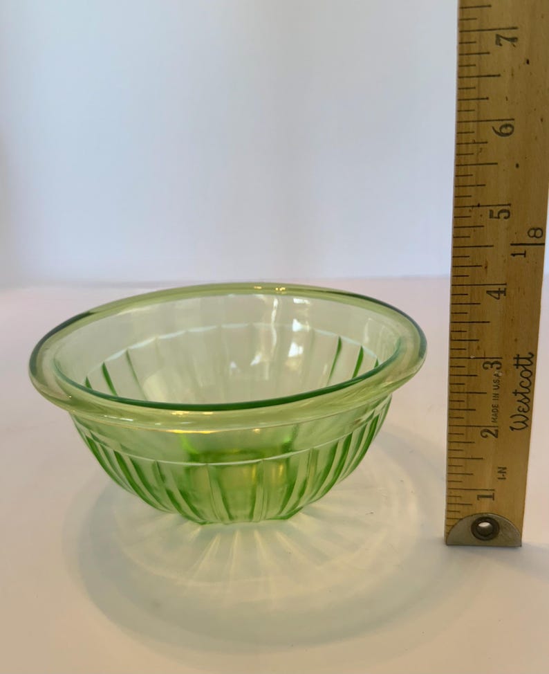May include: A small, light green glass bowl with vertical ribbed detailing. The bowl has a rounded shape with a slightly flared rim. A wooden ruler is positioned next to the bowl, showing measurements in inches.