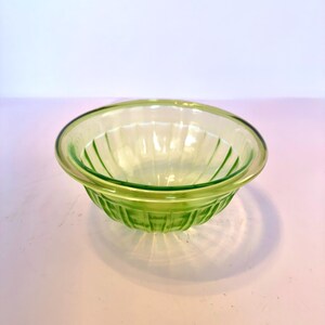 May include: A clear, green glass bowl with a ribbed design. The bowl has a rounded shape with a slightly flared rim. The glass has a subtle, light green hue, and the bowl is sitting on a white surface.