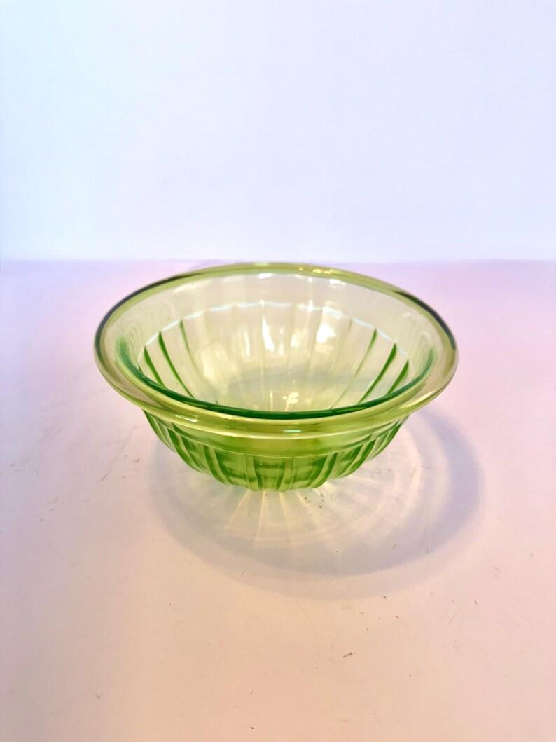 May include: A clear, green glass bowl with a ribbed design. The bowl has a rounded shape with a slightly flared rim. The glass has a subtle, light green hue, and the bowl is sitting on a white surface.