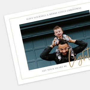 Op de afbeelding: Een vader en zoon zijn gefotografeerd voor een blauwe garagedeur. De vader draagt de zoon op zijn schouders. De zoon glimlacht en kijkt naar de camera. De tekst "HAVE YOURSELF A MERRY LITTLE CHRISTMAS" staat bovenaan de afbeelding. De tekst "LET YOUR HEART BE" staat onderaan de afbeelding. Het woord "light" is geschreven in gouden letters.