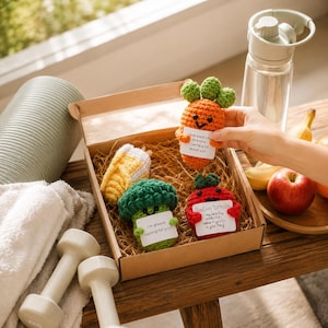 May include: A cardboard gift box filled with straw contains three crocheted vegetable figures: an orange carrot, a green broccoli, and a red tomato. Each figure holds a small white card with printed text. The carrot card reads: Just want you to know, I carrot a lot about you. The broccoli card reads: I'm always rooting for you! The tomato card reads: Positive tomato. I may be a tiny tomato but I believe in you. Go do your thing.