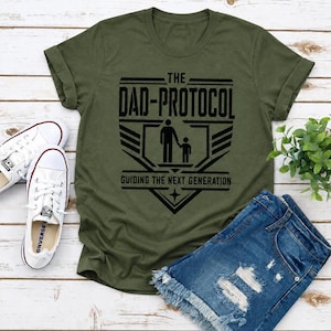 Op de afbeelding: Een olijfgroen T-shirt met korte mouwen en een zwarte grafische print met de tekst THE DAD-PROTOCOL GUIDING THE NEXT GENERATION, vergezeld van een gestileerd icoon van een volwassene die de hand van een kleiner kind vasthoudt.