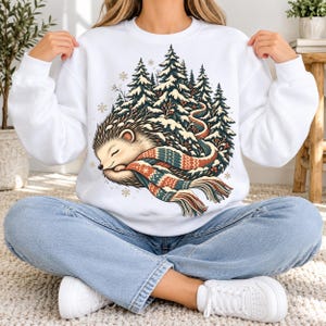 May include: A white crewneck sweatshirt featuring a graphic illustration of a sleeping hedgehog wrapped in a long, multi-colored patterned scarf. The hedgehog is positioned in front of a dense forest of snow-covered pine trees and snowflakes.