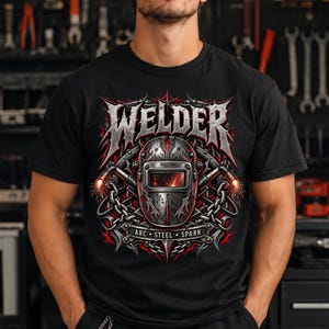 May include: A black t-shirt featuring a graphic print of a welding helmet surrounded by chains and two welding torches. The word WELDER is printed in large, metallic-style lettering above the helmet, with the words ARC, STEEL, SPARK below it.