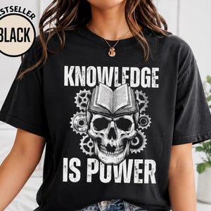 May include: A black short-sleeve t-shirt featuring a white graphic of a human skull with an open book resting on top, surrounded by mechanical gears. The text "KNOWLEDGE IS POWER" is printed in a distressed white font above and below the graphic.