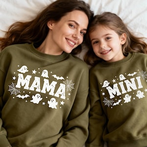 Può includere: Due felpe verde oliva con grafiche bianche a tema Halloween. Una felpa dice "MAMA" e l'altra "MINI". Il design include fantasmi, pipistrelli, ragnatele e stelle. Una madre e suo figlio indossano le felpe abbinate.