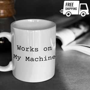 Peut inclure: Mug en céramique blanche avec l'inscription "Works on My Machine" en noir. La tasse a une anse incurvée et se détache sur un fond flou. Un graphique "Livraison GRATUITE" est visible.