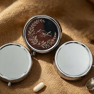 May include: Three silver-colored, round pill cases with a brown interior. One case has a floral design and the name "Krystina" in cursive. A small spoon and two pills are also visible on a textured, tan surface.