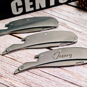 May include: Three silver-colored straight razors are displayed on a wooden surface. The razors have a curved design with a pointed tip and a rounded handle. One razor has the name "Tesory" engraved on it.