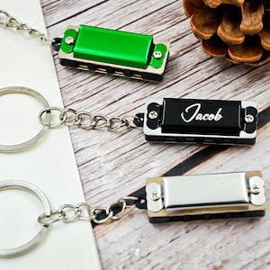 May include: Miniature harmonica keyrings in green, black with "Jacob" script, and silver. Each keyring features a small harmonica attached to a silver chain and ring. The keyrings are displayed on a wooden surface.
