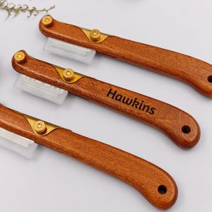 May include: Three wooden craft knives with brass accents and the name "Hawkins" engraved on one. The knives have a curved handle and a slot for a blade. The blades are protected by a clear plastic cover. The knives are arranged on a white surface.