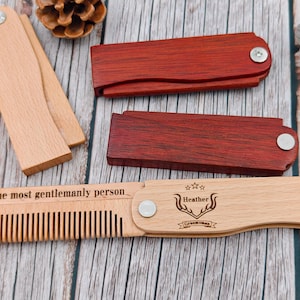 May include: Wooden folding combs in natural and reddish-brown tones are displayed on a wooden surface. One comb is engraved with "The most gentlemanly person" and a name. The combs are ideal for grooming and make a great gift.