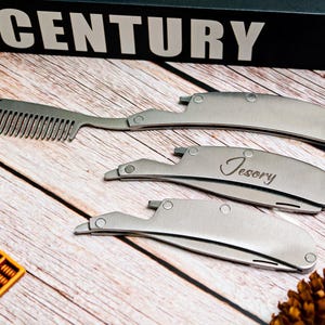 May include: Three silver folding combs and razors are arranged on a wooden surface. One comb is open, while the other two are closed. The word "Century" is visible on a black box in the background. One razor is engraved with the word "Tesory".