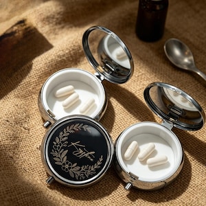 May include: Three open, round, silver pill cases with mirrors, each containing white capsules. One case has a black lid with a floral design and the name "Kristina". A small bottle and spoon are also visible.