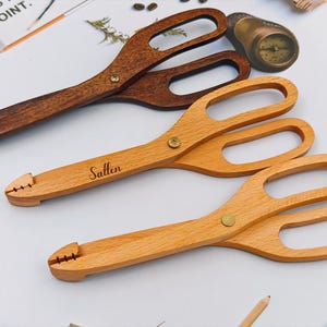 Puede incluir: Tres pinzas de servir de madera en tonos marrones y beige. La pinza superior es de color marrón oscuro, la del medio es de color marrón claro con el nombre "Sallen" grabado, y la inferior es de color beige claro. Cada una tiene un mango en forma de tijera y una cabeza ranurada.