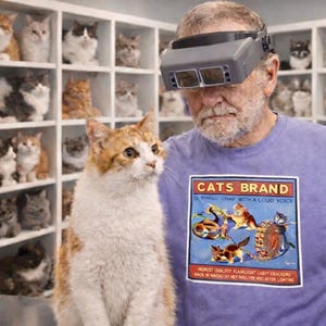 May include: A ginger and white cat sits in front of a man wearing a purple t-shirt with a "CATS BRAND" graphic. The man wears a head-mounted magnifier. Shelves filled with cat figurines are in the background.