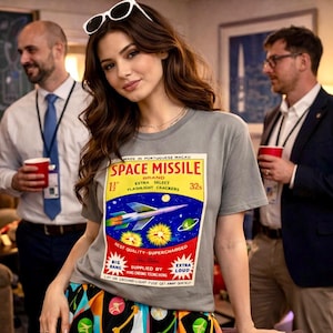 May include: A gray t-shirt with a colorful graphic of a space missile, stars, and planets. The text on the shirt reads "SPACE MISSILE" and "EXTRA SELECT FLASHLIGHT CRACKERS". The shirt is worn by a person in a room with other people.