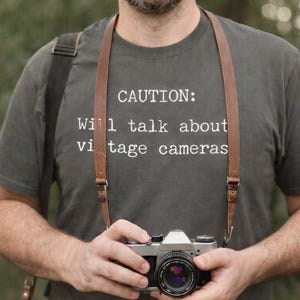 May include: A gray t-shirt with the text "CAUTION: Will talk about vintage cameras." A vintage camera with a brown leather strap is held in front of the shirt. The camera has a black lens and silver body.