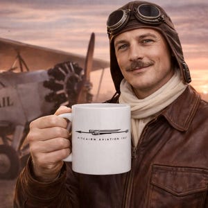 May include: White ceramic mug with the text "Pitcairn Aviation 1927" and a stylized airplane design. The mug is held in front of a vintage airplane and a person wearing a leather aviator jacket and goggles.