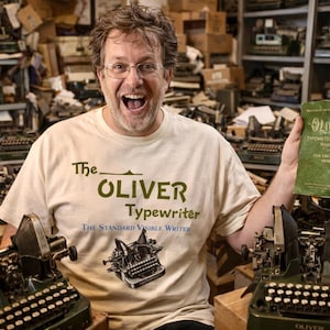 May include: A man wearing glasses and a cream-colored t-shirt with "The Oliver Typewriter" logo, holding a green manual. The background is filled with vintage typewriters, suggesting a collection or workshop.