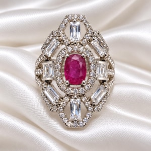 May include: A silver-toned ring featuring a central oval-cut deep pink gemstone surrounded by a halo of small round clear stones. The frame is accented with six rectangular baguette-cut clear stones arranged in a geometric pattern.
