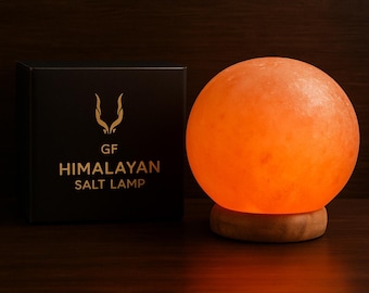 May include: A glowing Himalayan salt lamp with a round, orange-hued salt crystal sits on a wooden base. A black box with gold lettering that reads "GF HIMALAYAN SALT LAMP" is next to the lamp. The background is a dark wood surface.