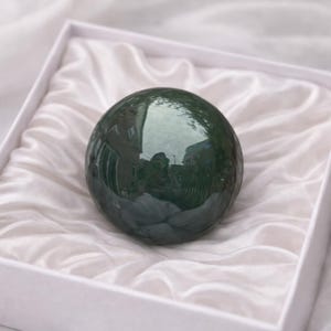 May include: A dark green, polished sphere rests in a white box lined with white satin fabric. The sphere reflects the surrounding environment. The box is square and has a simple design. The overall presentation suggests a luxury item or a decorative object.