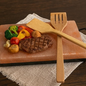 Puede incluir: Un bloque de sal rosa del Himalaya con un bistec a la parrilla y verduras coloridas, servido con espátulas de madera. El bloque de sal está sobre un marco de metal negro, sobre una servilleta de lino. Una presentación culinaria.