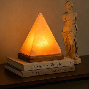 Puede incluir: Lámpara de sal del Himalaya en forma de pirámide con un brillo naranja cálido, sobre una base de madera. La lámpara está sobre dos libros con los títulos "The Art of Simple" y "Walter Isackson". Una estatua clásica en el fondo.