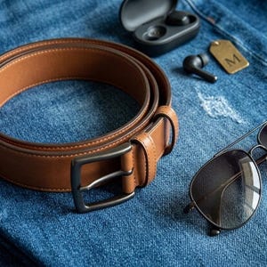 Pode incluir: Um cinto de couro castanho com fivela de metal escuro, óculos de sol estilo aviador com lentes escuras e armação preta, um estojo de carregamento preto para auriculares sem fios, um auricular solto e uma etiqueta de latão com a letra M.