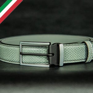 Puede incluir: Un cinturón verde claro con un patrón de piel de serpiente. El cinturón tiene una hebilla plateada y una barra negra. La bandera italiana está en la esquina superior izquierda. El cinturón está sobre una superficie negra.