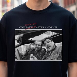 Könnte beinhalten: Schwarzes T-Shirt mit einem Schwarz-Weiß-Bild von zwei Männern in einem Auto, mit dem Text "Clock-Out" und "ONE BATTLE AFTER ANOTHER" in Rot und Weiß. Das Bild zeigt zwei Männer, von denen einer eine Dose hält.