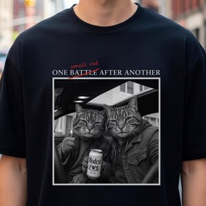 Könnte beinhalten: Schwarzes T-Shirt mit einem Schwarz-Weiß-Bild von zwei Katzen in einem Auto. Eine Katze hält ein brennendes Streichholz, die andere eine Dose "Modecat"-Bier. Der Text über dem Bild lautet "ONE BATTLE AFTER ANOTHER".