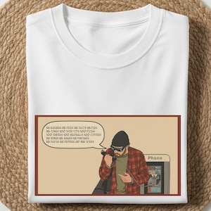 May include: White t-shirt with a graphic of a person in a red plaid shirt talking on a payphone. The graphic includes a speech bubble listing food items like burgers, fries, tacos, and pies. The word "Phone" is also visible.