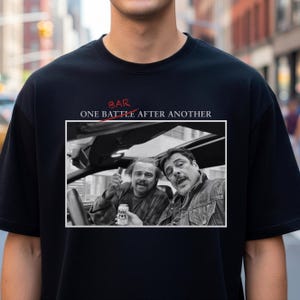 Könnte beinhalten: Schwarzes T-Shirt mit einem Schwarz-Weiß-Bild von zwei Männern in einem Auto und dem Text "ONE BATTLE AFTER ANOTHER" über dem Bild. Das Wort "BAR" steht über dem Text, wobei das "R" rot durchgestrichen ist.
