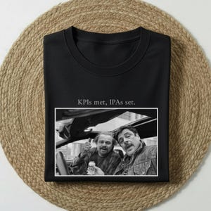 Könnte beinhalten: Schwarzes Sweatshirt mit Rundhalsausschnitt und dem Text "KPIs met, IPAs set." Auf der Vorderseite des Sweatshirts ist ein Schwarz-Weiß-Foto abgebildet. Das Foto zeigt zwei Männer in einem Auto, einer hält eine Bierdose.