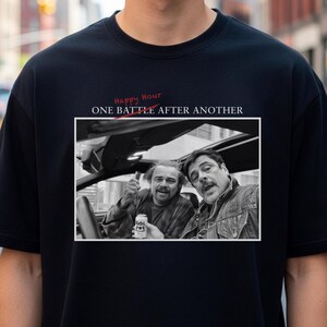 May include: Black t-shirt featuring a black and white image of two men in a car. The image is framed with the text "ONE BATTLE AFTER ANOTHER" and "Happy Hour" in red script.