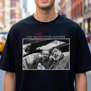 Könnte beinhalten: Schwarzes T-Shirt mit einem Schwarz-Weiß-Bild von zwei Männern in einem Auto, einer hält eine Bierdose. Der Text "BAR" und "ONE BATTLE AFTER ANOTHER" sind in Rot und Weiß über dem Bild gedruckt.