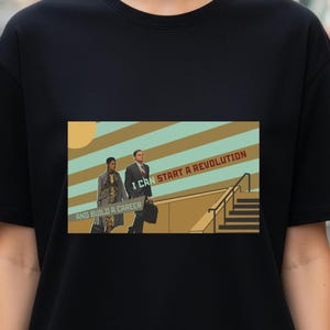 May include: Black t-shirt featuring a graphic with two people walking, with the text "I CAN START A REVOLUTION AND BUILD A CAREER" in a retro style. The design includes a sun and a staircase.