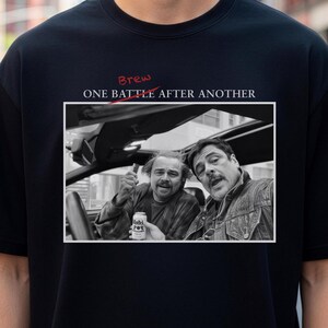 Könnte beinhalten: Schwarzes T-Shirt mit einem Schwarz-Weiß-Bild von zwei Männern in einem Auto und dem Text "ONE BATTLE AFTER ANOTHER". Das Wort "Brew" ist rot durchgestrichen.