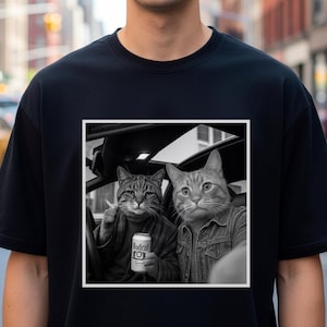 Könnte beinhalten: Schwarzes T-Shirt mit einem Schwarz-Weiß-Bild von zwei Katzen in einem Auto. Eine Katze hält eine Dose "Modeal Toy Special", die andere trägt eine Jeansjacke. Das Bild ist in einem weißen Quadrat gerahmt.