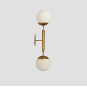 Op de afbeelding: Een wandlamp met een verticale, messingkleurige metalen stang verbonden aan een ronde muurplaat. De armatuur bevat twee bolvormige witte glazen bollen, één aan de bovenkant en één aan de onderkant van de centrale stang geplaatst.